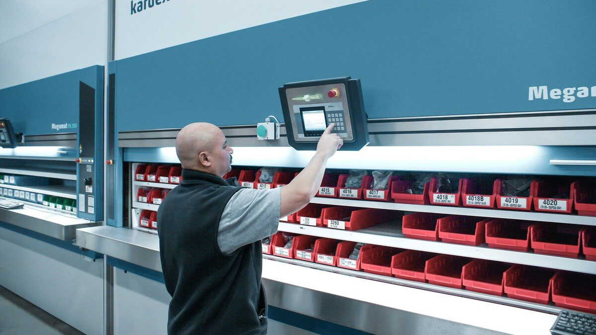 Warehouse operator retrieving items from a Kardex Remstar vertical carousel storage system using a touchscreen interface with red bin compartments