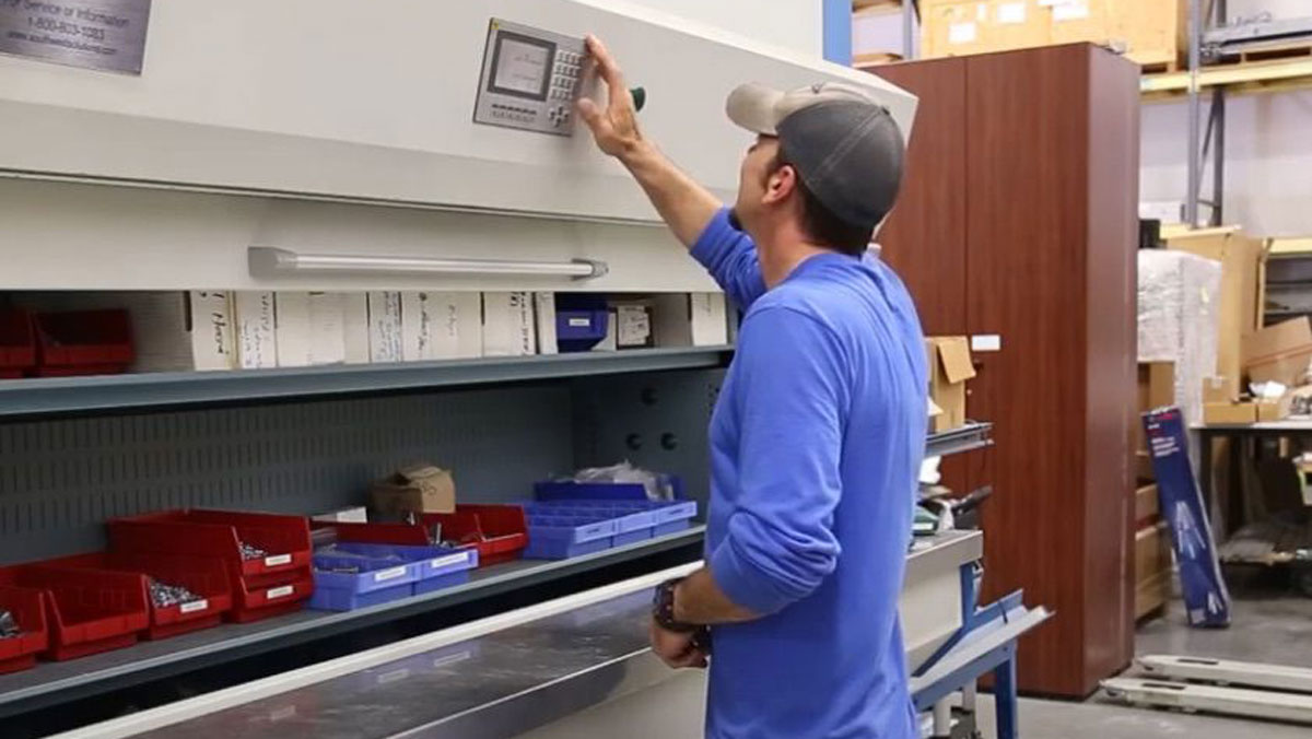 Warehouse worker operating a vertical carousel storage unit manually using a control panel with red and blue storage bins visible on the tray