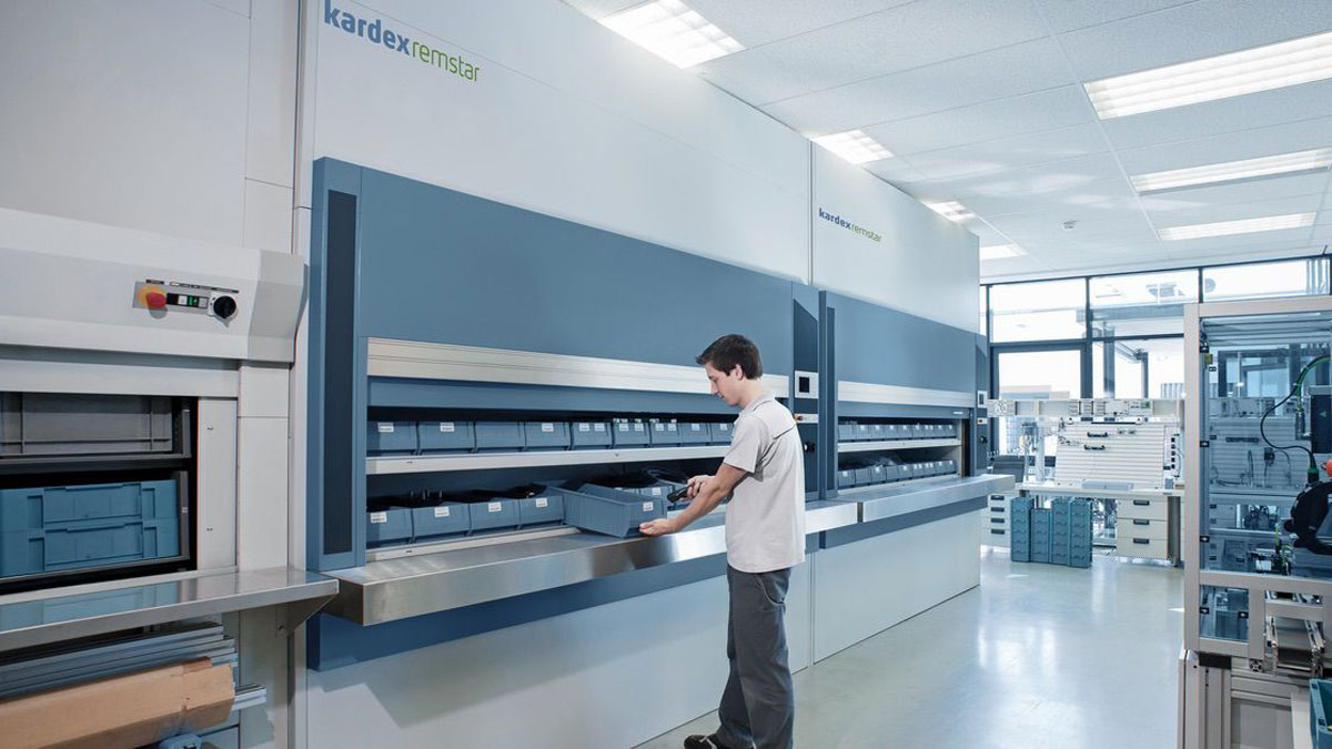 Technician organizing blue storage bins inside a Kardex Remstar vertical carousel system with a well-lit workspace and high-ceiling lab