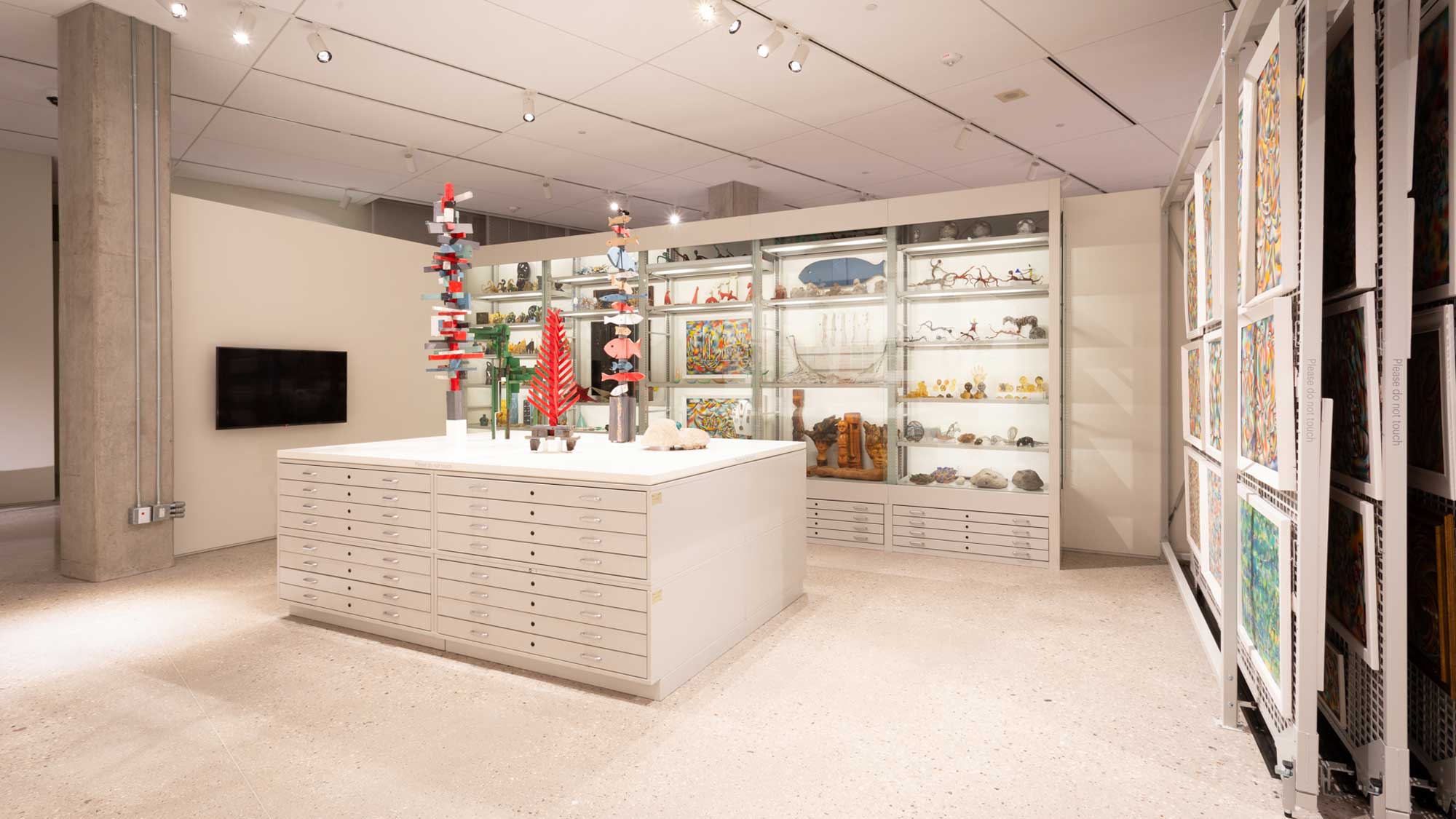 Conservation storage area with flat-file drawers below illuminated museum display cases holding art objects.