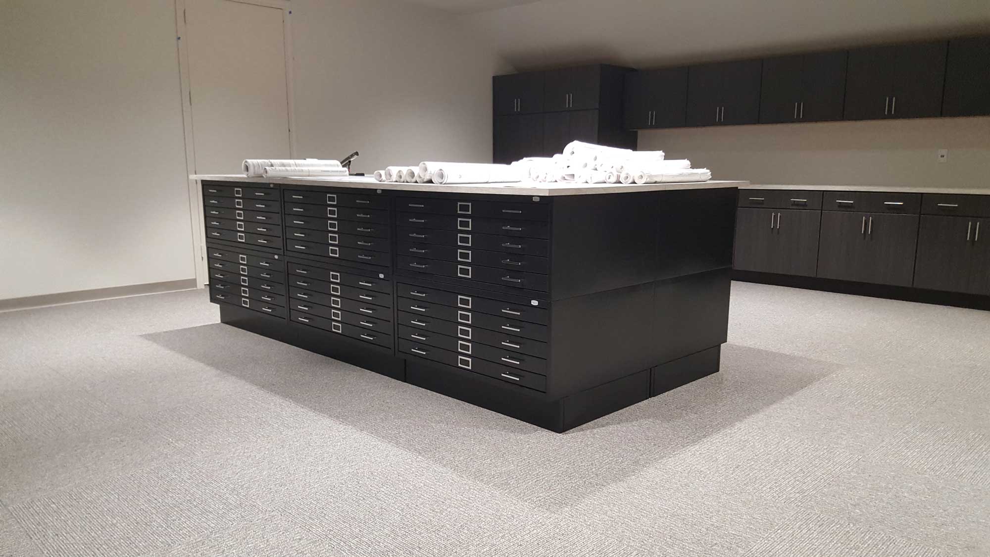 Architectural flat-file island with black pull-out drawers and white countertop holding rolled blueprints in design studio.