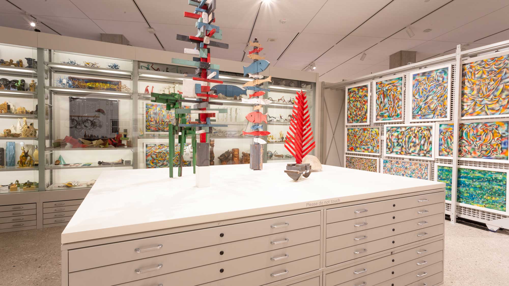 Central flat-file display island topped with sculptures, flanked by vertical sliding racks of framed artworks in gallery.