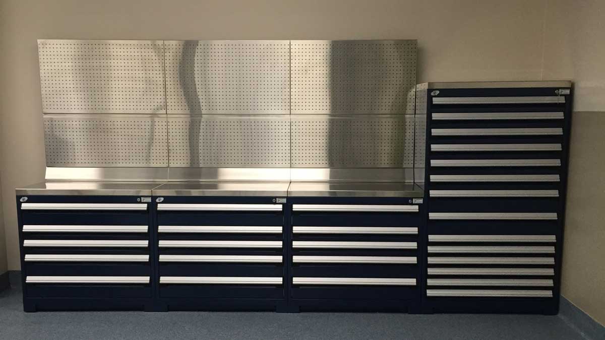 Black modular drawer cabinets topped by a stainless steel countertop and perforated steel pegboard. 