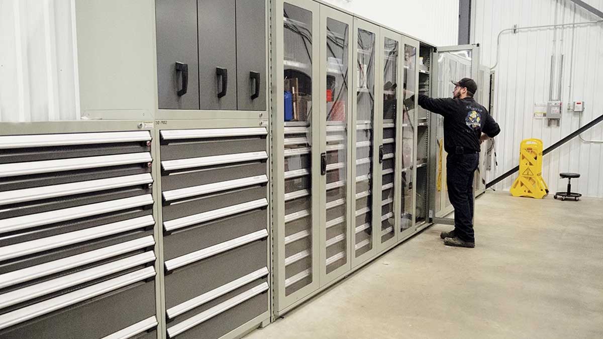 Combination of grey drawer cabinets and upper lockable cabinets, with a technician retrieving parts.