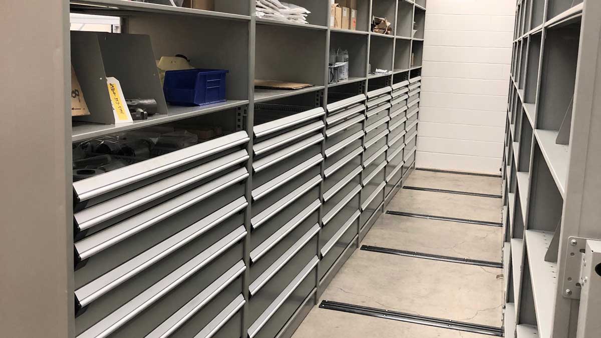 Grey modular drawer cabinets lining a narrow aisle between shelving units in a parts storeroom. 