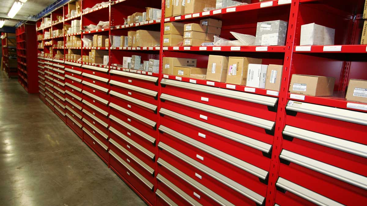 Rows of red modular drawer cabinets with upper shelves holding cardboard boxes in a warehouse corridor. 