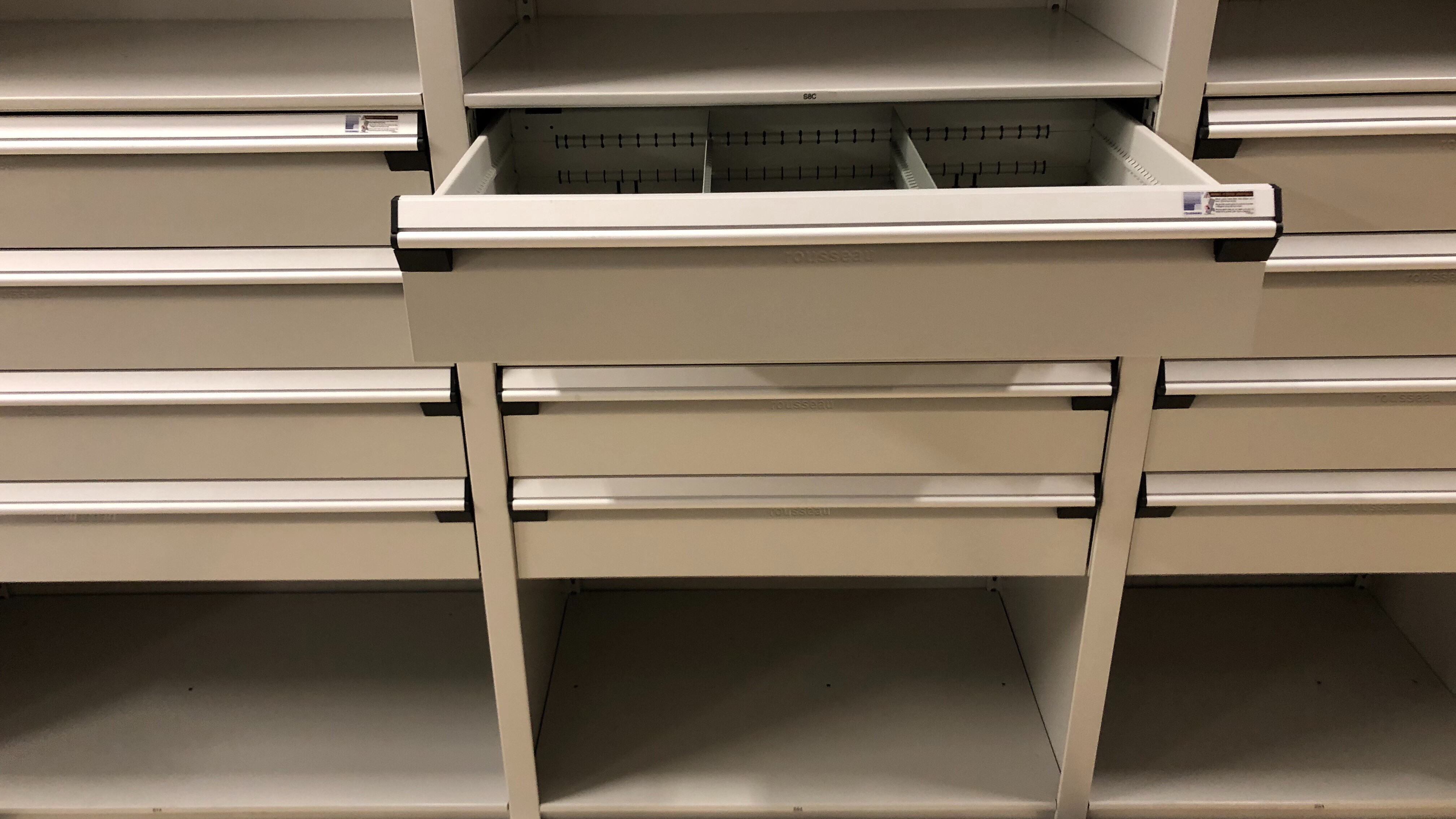 Front-facing tan modular drawer cabinet with one open drawer showing divider compartments under upper shelves.