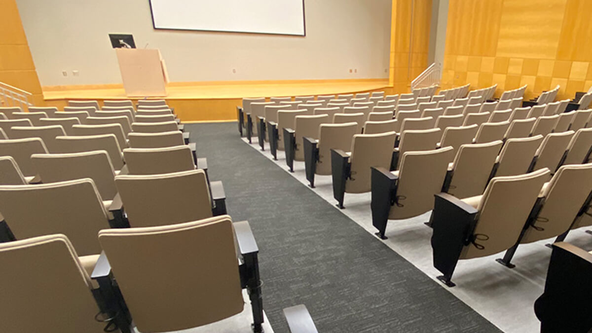 lecture hall seating featuring beige upholstered chairs in tiered rows facing a stage and presentation screen
