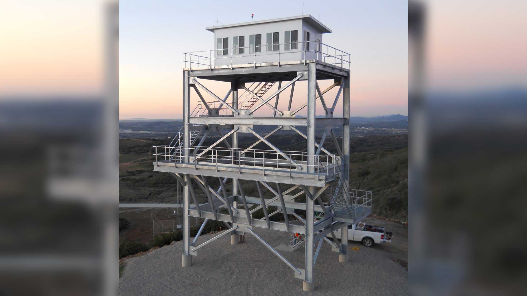 galvanized steel multi-level observation tower with two wraparound platforms and an enclosed top cabin at dusk overlooking a valley 