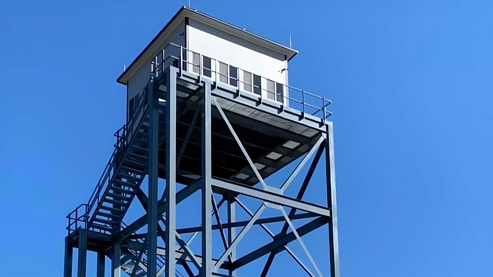 galvanized steel multi-level training tower featuring open staircases, wraparound platforms with yellow guardrails, and a sheltered top cabin against a blue sky
