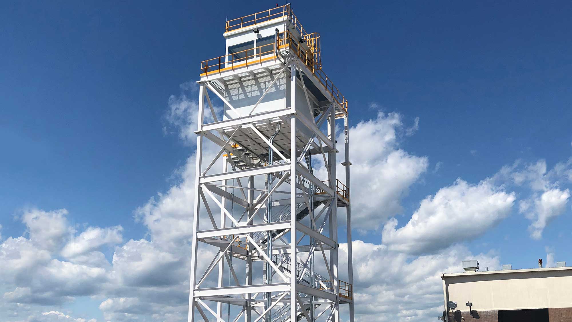 tall white steel observation tower with multiple grated platforms and bright yellow safety railings, topped by an enclosed cabin under a clear blue sky