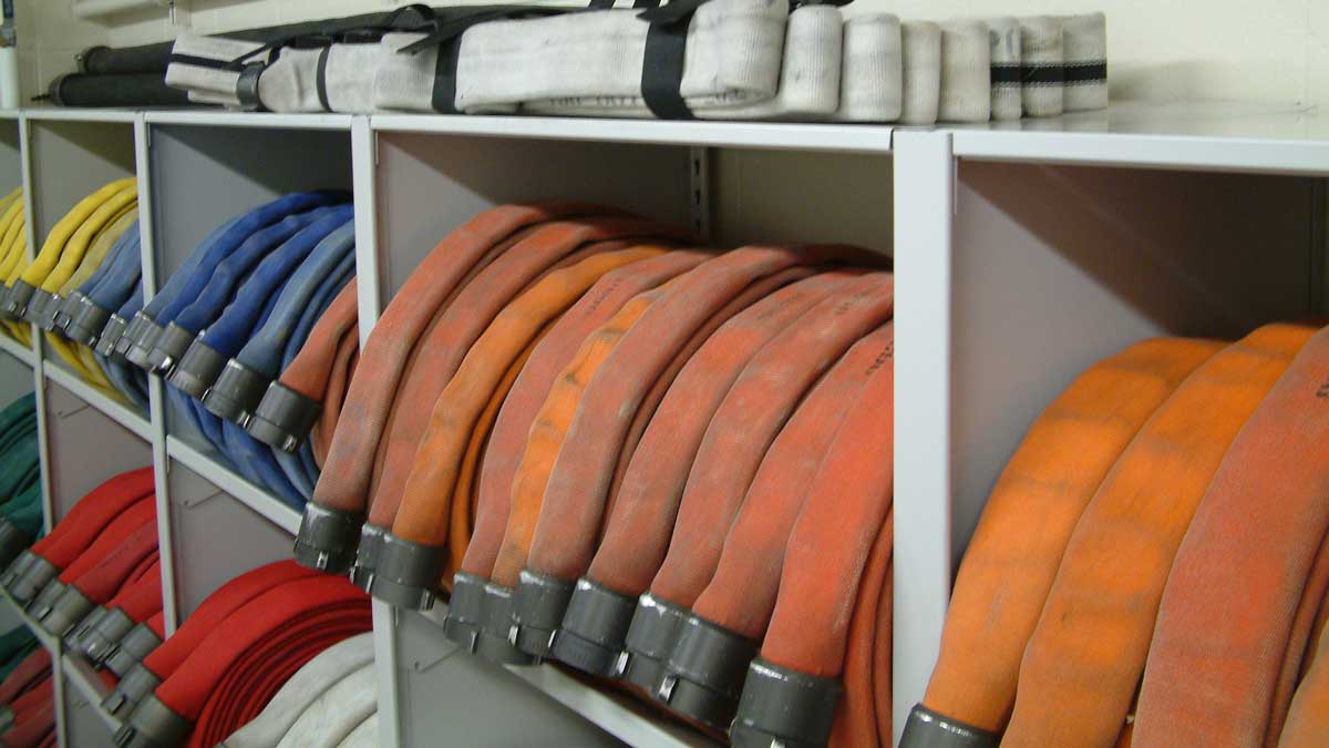 Color-coded fire hoses neatly rolled and stored in individual compartments on a metal shelving unit in a fire department equipment room.