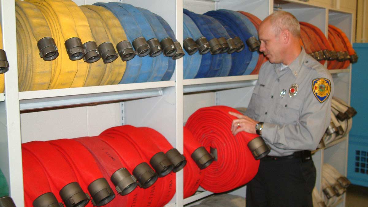 Fire department officer reaching for a bright red rolled fire hose on a heavy-duty metal shelf stocked with yellow, blue, and orange hoses.