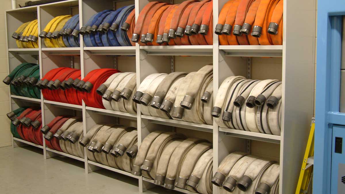 Wide view of multiple rows of coiled fire hoses organized by color on adjustable white shelving in a fire station.