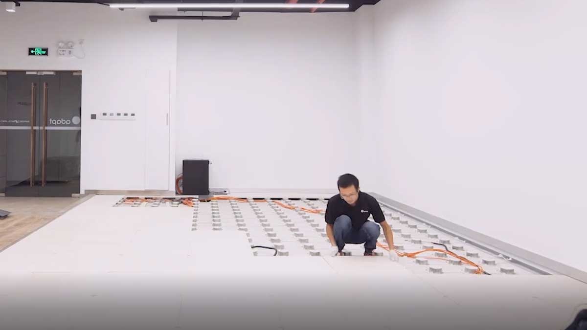 A technician kneels on an unfinished raised-floor grid, installing floor panels over adjustable pedestals. The room is a bright, white office space with electrical and data cables visible beneath the panels.